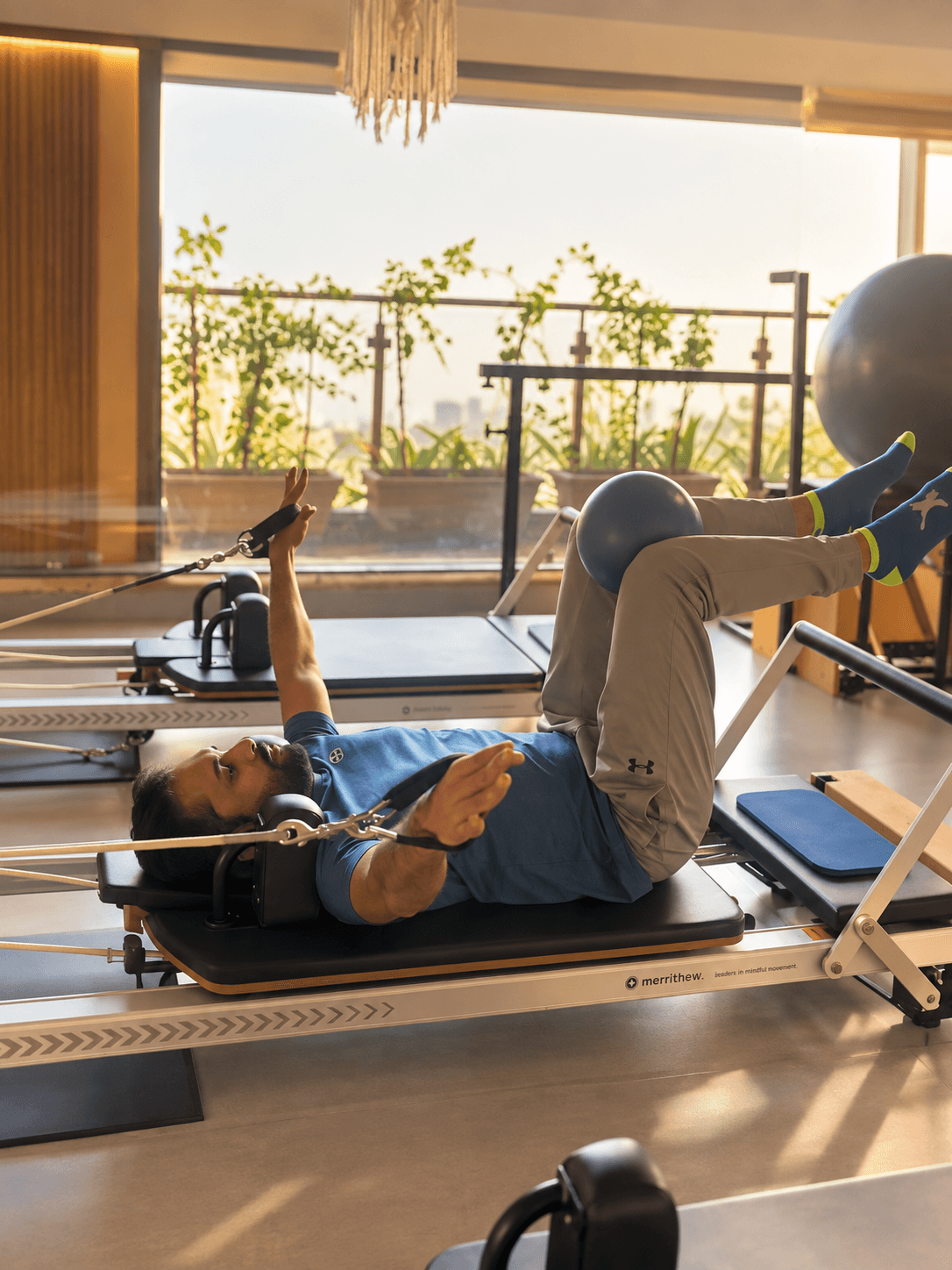 A man on a Pilates reformer, one leg raised against a small exercise ball and both arms reaching overhead on the straps, the studio's floor-to-ceiling windows and skyline behind him.