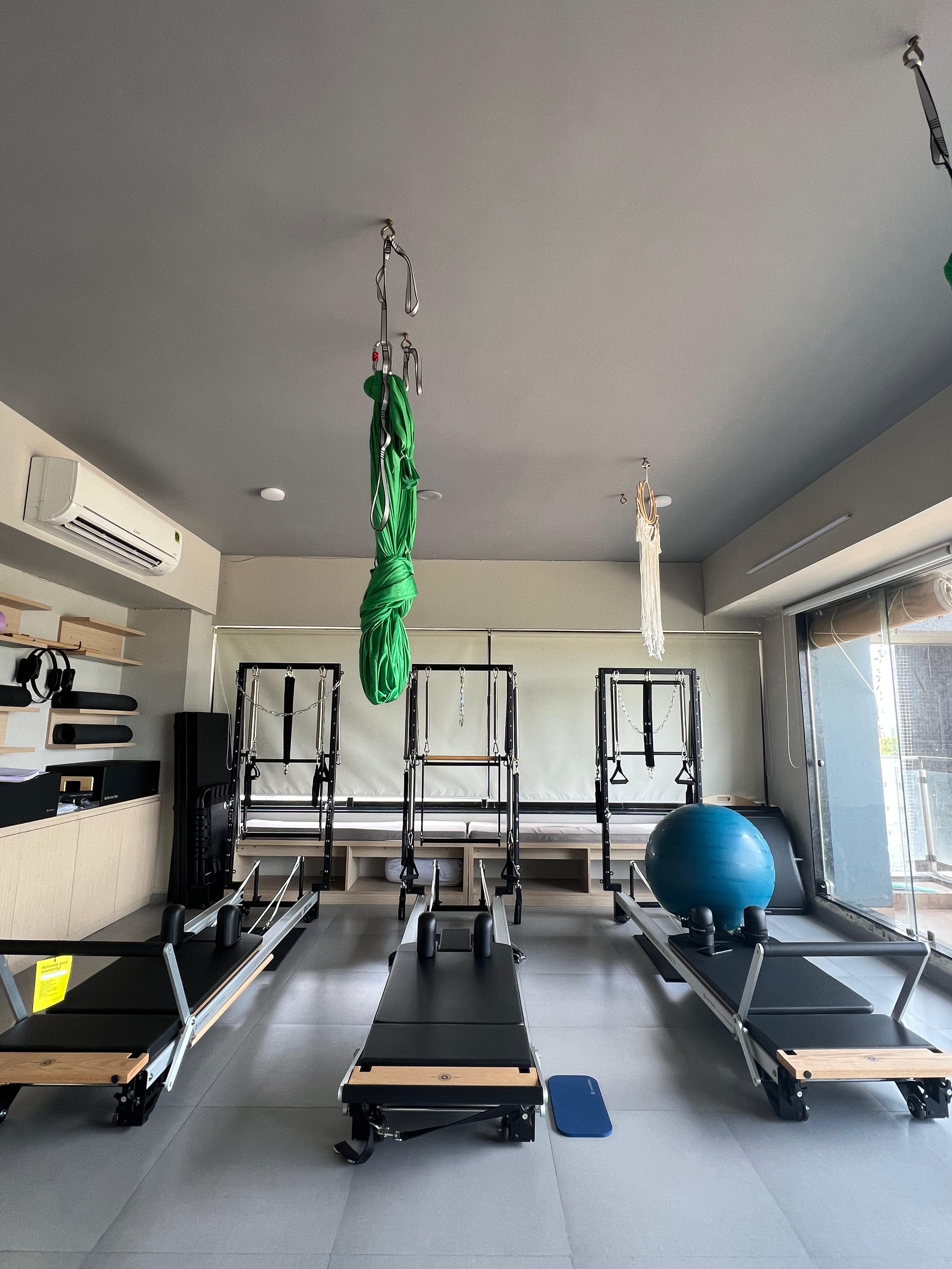 The studio's three reformers in a row facing the floor-to-ceiling window, a blue exercise ball and shelving of folded towels to the side, green aerial silks hanging from the ceiling above.